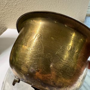 Può includere: Un vaso per piante rotondo in ottone con finitura metallica lucida, caratterizzato da disegni a linee incise raffiguranti un uccello appollaiato su un ramo e fogliame circostante. Il vaso ha un bordo largo e poggia su tre piccoli piedini curvi.