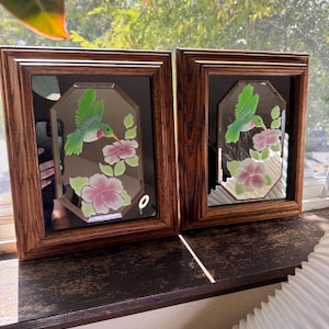 May include: Two rectangular wall mirrors with dark brown wood-grain frames. Each mirror features an octagonal center panel with a painted design of a green hummingbird hovering over pink flowers with green leaves against a reflective surface.