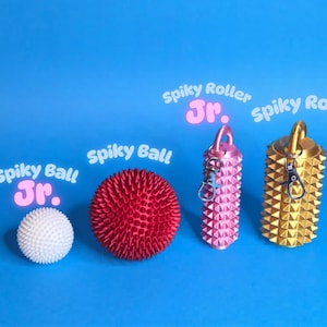 May include: A collection of spiky massage tools and toys on a blue background. The items include a white spiky ball, a red spiky ball, a pink spiky roller, and a gold spiky roller. Text on the image reads "Spiky Ball Jr." and "Spiky Roller Jr."