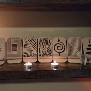 May include: A collection of seven wooden phone stands with various cut-out designs, including a fern, geometric patterns, and a spiral. The stands are displayed on a dark wooden shelf, with two lit tea light candles in front.