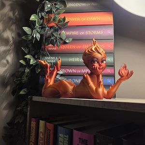 May include: A bronze-colored figurine of a reclining woman with a flame-shaped head and a small flame in her hand. The figurine is resting on a shelf in front of a stack of books with colorful spines. Green vines are draped on the side.