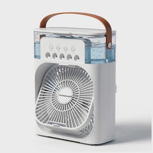 Puede incluir: Un enfriador de aire portátil blanco con un depósito de agua transparente con gotas visibles, una rejilla de ventilador circular y un asa de transporte de cuero marrón en la parte superior. El panel de control incluye cuatro botones circulares.