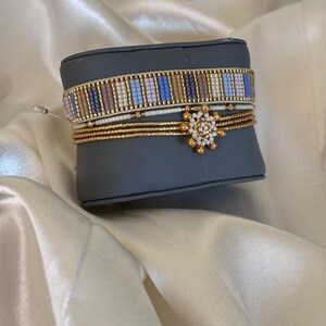 May include: A collection of beaded bracelets in various styles. One bracelet features a striped pattern in blue, purple, and tan. Other bracelets are gold and white with a gold floral accent. The bracelets are displayed on a gray surface.