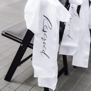 Op de afbeelding: Zwarte klapstoel met een witte sjerp over de rug. Op de sjerp staat het woord "Reserved" in zwart schrift. De stoelen staan op een grijs metalen oppervlak, geschikt voor evenementen.