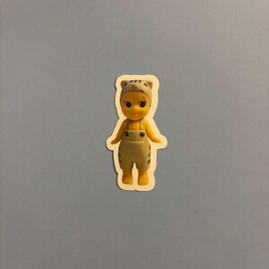 Puede incluir: Una pequeña figura amarilla que lleva un gorro color canela con rayas oscuras y un peto a juego del mismo color con rayas oscuras. La figura tiene la cabeza redonda, ojos grandes y oscuros y una pequeña nariz rosa. Es una pegatina troquelada.