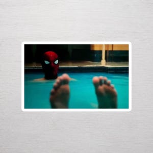 May include: A sticker featuring a red and black mask with large white eye lenses, partially submerged in bright blue swimming pool water, with two feet visible in the foreground above the water surface, creating a humorous and cinematic scene.