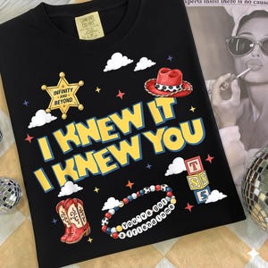 May include: A black t-shirt featuring a graphic print with the text "I KNEW IT I KNEW YOU" in yellow. The design includes a sheriff star badge with "INFINITY AND BEYOND", a red cowboy hat, cowboy boots, a beaded bracelet with "YOU'VE GOT A FRIEND IN ME", and blocks.