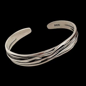 May include: A silver cuff bracelet with a textured, wave-like design. The open-ended bracelet has a smooth, polished finish on the inner band and the text "S925" is visible. The bracelet is displayed against a black background.