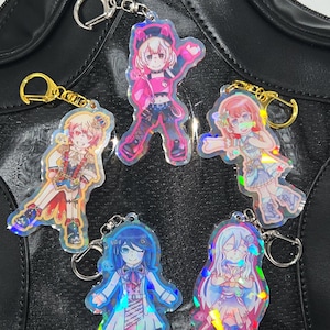 May include: Five anime-style character keychains with silver and gold-colored hardware. Each keychain features a different character design with vibrant colors and holographic accents. The keychains are attached to a black leather bag.