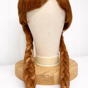 May include: A red-brown wig with two long, braided pigtails. The wig is styled on a mannequin head, with a light-colored section at the crown. The braids are secured at the ends. The wig is displayed on a wooden stand.