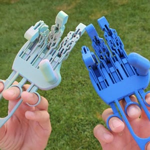 May include: Two 3D-printed robotic hands, one light blue and one dark blue, are held up against a green grassy background. The hands are detailed with articulated fingers and a mechanical design.
