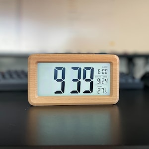 May include: A rectangular digital clock with a light wood frame displays the time as 9:39. The screen also shows the alarm time, date, and temperature of 27.5 degrees Celsius. The clock sits on a dark surface.