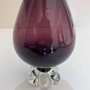 May include: A deep purple glass vase with a wide, rounded body that tapers towards the top opening. The vase is supported by a clear, thick glass base featuring a distinct, multi-lobed, petal-like design that provides a stable foundation.