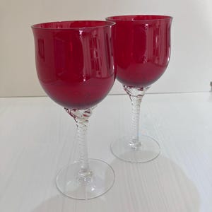 May include: Two wine glasses with deep red bowls and clear, twisted glass stems. Each glass features a wide, rounded bowl shape and a circular clear glass base. The stems have a distinct spiral pattern that connects the bowl to the base.