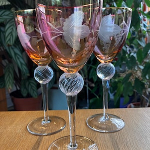 May include: Three vintage wine glasses with pink and gold rims. Each glass features a clear, etched grape and leaf design. The stems have a decorative ball and swirl design, and the bases are round.