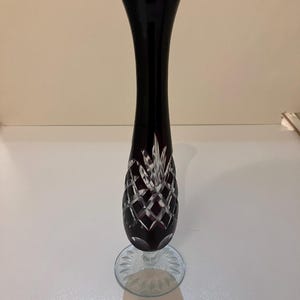 May include: A tall, slender bud vase crafted from deep purple glass. The base of the vase features a clear glass pedestal with a starburst pattern, while the body displays a cut-glass diamond lattice design that reveals the clear glass beneath.