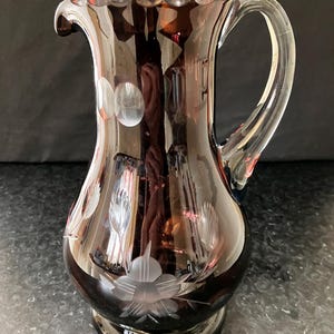 May include: A dark amber glass jug with a clear glass handle. The jug features etched floral and dot designs. The jug is approximately 23 cm tall.