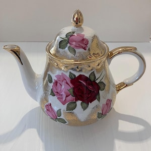 May include: A white porcelain teapot featuring a floral pattern of pink and dark red roses with green leaves. The teapot is accented with gold trim on the spout, handle, lid finial, and along the upper rim, with gold sponged detailing.
