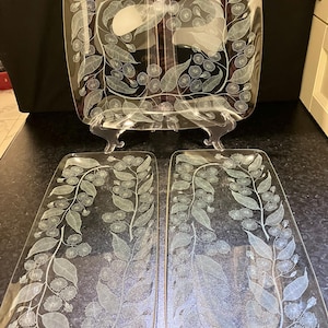 May include: A set of three clear glass serving platters featuring a white etched botanical pattern of eucalyptus leaves and blossoms. One large square platter is displayed upright on a stand, while two rectangular platters lie flat below it.
