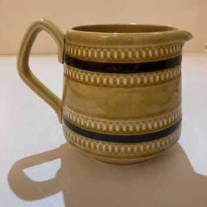 May include: A mustard yellow ceramic jug featuring a glossy finish, a single handle, and two horizontal dark brown bands. The body is decorated with embossed, repeating oval patterns that border the dark brown stripes on the upper and lower sections.