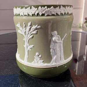 May include: A sage green ceramic planter featuring white relief decorations of a classical figure standing by a pillar, a tall tree, and a leafy vine border around the top rim. The cylindrical vessel has a smooth, glossy finish.