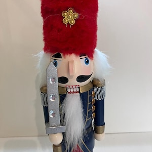 May include: A wooden nutcracker figurine wearing a tall red fuzzy hat with a gold flower emblem. The figure has a white faux fur beard, a blue uniform with silver fringe epaulets, and holds a silver sword decorated with three clear rhinestones.