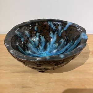 May include: A round ceramic bowl with a dark brown exterior featuring a textured, pebble-like surface. The interior of the bowl has a glossy finish with a deep brown base colour and vibrant, flowing streaks of bright blue glaze radiating outwards.