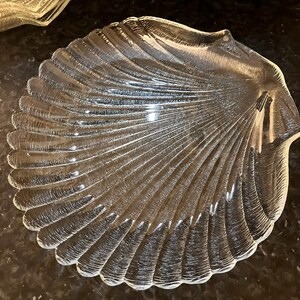May include: Clear glass seashell-shaped serving dish. The dish has a detailed, ridged design that mimics the texture of a seashell. The dish is transparent and appears to be made of glass. The dish is likely used for serving food or as a decorative item.