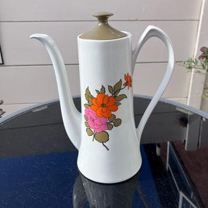 May include: A tall white ceramic coffee pot featuring a light brown lid and a floral graphic on the front. The design displays orange and pink flowers with olive green leaves and stems. The white body has a subtle crackle glaze finish.