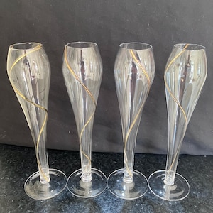 May include: A set of four clear glass champagne flutes, each featuring a single delicate gold-coloured spiral line wrapping around the exterior of the bowl. The glasses have a slender, elongated shape with a circular base and a smooth, transparent finish.