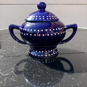 May include: A deep blue ceramic sugar bowl with a matching lid, featuring two curved handles and a pedestal base. The bowl and lid are decorated with horizontal rows of small, white painted dots encircling the body and the base.