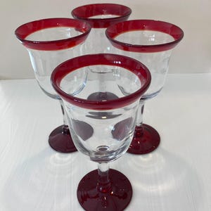 May include: A set of four clear glass goblets, each featuring a deep red rim and a matching deep red circular base. The stems are clear, connecting the bowl to the base, with a consistent design across all four pieces of glassware.