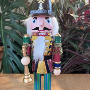 May include: A wooden nutcracker figure wearing a black helmet with gold wheat detailing, a red jacket with gold accents, and green and blue striped trousers. The figure has a white faux fur beard, a black moustache, and holds a gold trumpet.