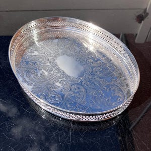 May include: An oval silver-toned serving tray featuring an intricate embossed floral and scrollwork pattern across the base. The tray has a raised, openwork lattice border around the perimeter and a smooth, unadorned oval centre for engraving.