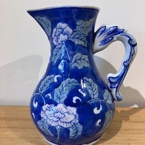 May include: A ceramic jug featuring a deep blue and white floral pattern with leafy vines. The jug has a curved handle with a sculpted, leaf-like design at the top and a wide pouring spout. The surface is glossy and smooth.