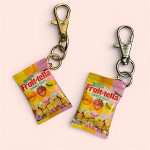May include: Two keychains with miniature Fruit-tella candy bags. The bags are yellow and pink, featuring images of fruit and the text "Fruit-tella Summer Fruits". One keychain has a gold clasp, the other a silver clasp.