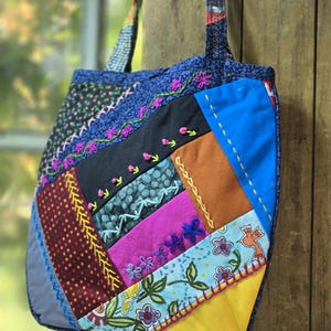 Peut inclure: Un sac fourre-tout en patchwork composé de divers morceaux de tissu bleu, jaune, noir, rose et marron. Le sac est orné de broderies colorées faites à la main, incluant des motifs floraux, des vignes et des lignes géométriques sur les panneaux.