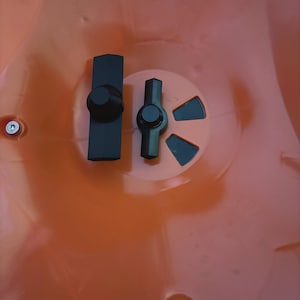 May include: Close-up of an orange plastic object with two black plastic components. One component is a rectangular shape with a circular knob, and the other is a T-shaped piece with a circular knob. Metal screws are visible.