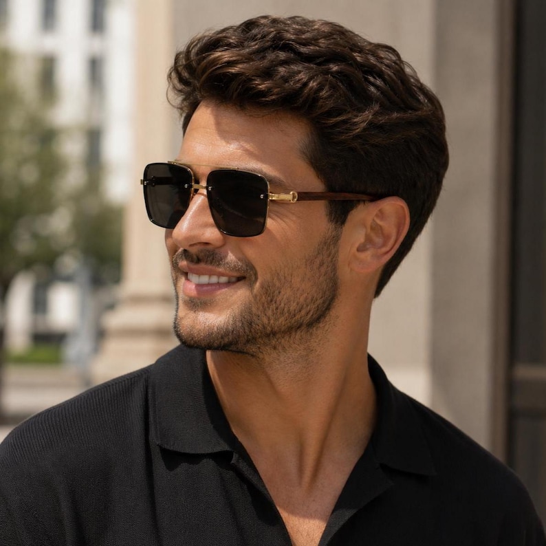 May include: Square-framed sunglasses with dark lenses and gold accents. The sunglasses have a brown wooden arm and are worn by a person wearing a black shirt. The person is smiling.