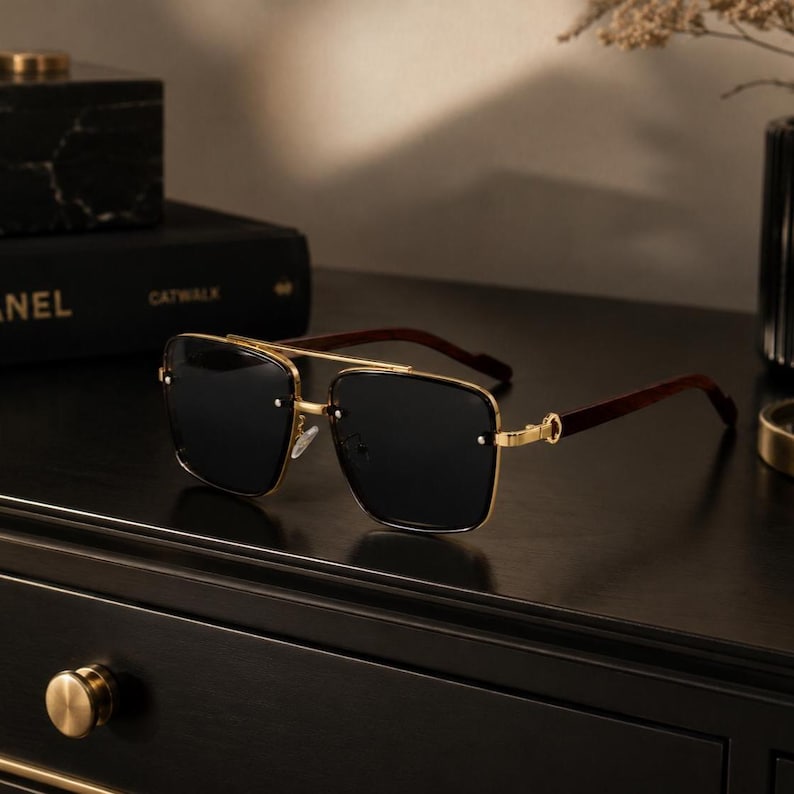 May include: Gold-framed sunglasses with dark lenses and brown wooden arms. The sunglasses have a double bridge and square lenses. The glasses are resting on a dark surface.