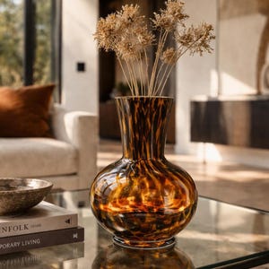 Peut inclure: Un vase décoratif en verre avec une base arrondie et un col évasé. Le vase présente un motif écaille de tortue dans des tons bruns et ambrés. Il contient des tiges beiges séchées et est posé sur une table en verre, suggérant un thème de décoration intérieure.