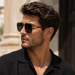 May include: Square-framed sunglasses with dark lenses, gold accents, and brown temples. The sunglasses have a double bridge and are worn by a person wearing a black shirt. The sunglasses are a stylish accessory.