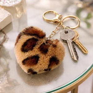 Peut inclure: Un porte-clés en forme de cœur avec un motif léopard duveteux dans les tons marron et beige. Le porte-clés est doté d'un anneau et d'une chaîne dorés, avec deux clés attachées. Les clés sont argentées et dorées.