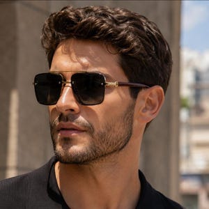 May include: Square-framed sunglasses with a gold-tone metal bridge and dark lenses. The temples are a dark brown color. The sunglasses are worn by a person.