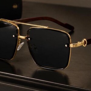 May include: Gold-rimmed sunglasses with dark lenses and brown wooden temples. The square frames have a double bridge and a classic, sophisticated design. The sunglasses are a stylish accessory.