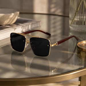 May include: Square-framed sunglasses with a gold metal frame and dark lenses. The temples are a dark wood grain. The sunglasses are resting on a reflective surface.