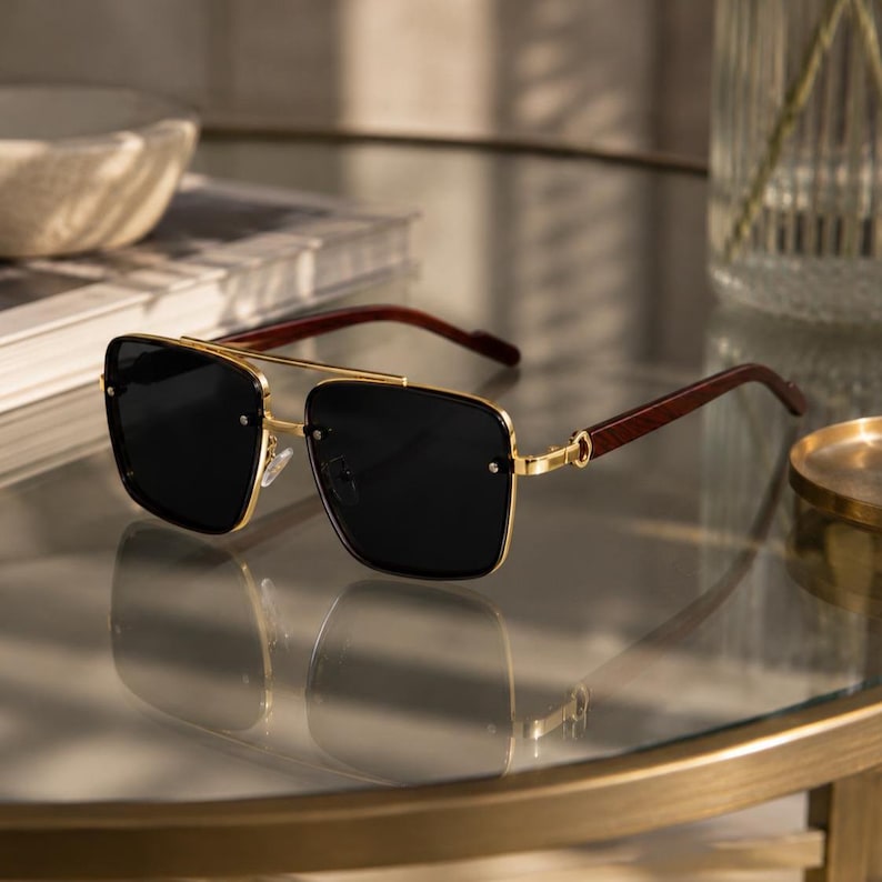 May include: Square-framed sunglasses with a gold metal frame and dark lenses. The temples are a dark wood grain. The sunglasses are resting on a reflective surface.
