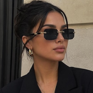 Könnte beinhalten: Schwarze rechteckige Sonnenbrille mit goldenen Akzenten. Die Sonnenbrille hat dunkle Gläser und wird von einer Person getragen. Der Rahmen ist schwarz mit goldenen Details an den Bügeln. Die Person trägt einen schwarzen Blazer und goldene Creolen.