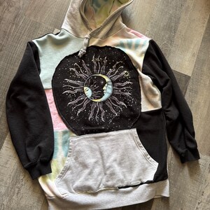 May include: A patchwork hoodie with a celestial design. The black sleeves and a large circular patch featuring a sun and moon graphic contrast with the tie-dye and pastel panels. A kangaroo pocket and hood complete the design.