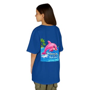May include: A royal blue t-shirt featuring a pink dolphin leaping out of the ocean with palm trees in the background. The text "Choose Joy & Make Waves" is printed below the dolphin. The shirt is worn by a person.